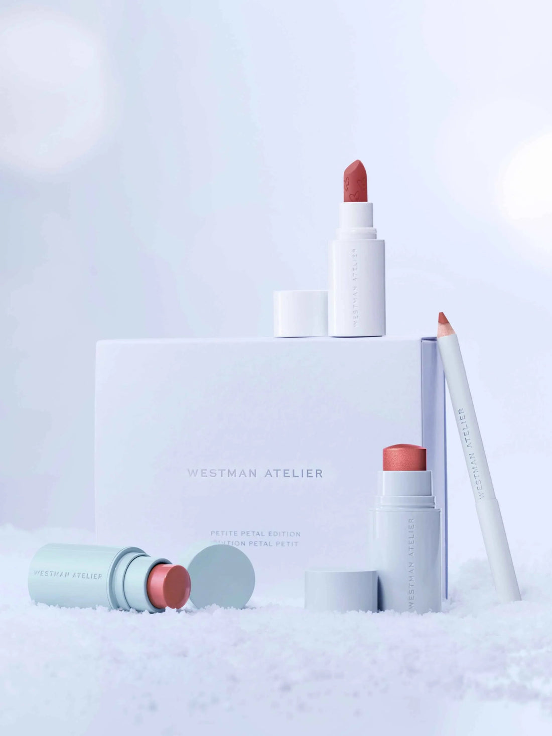 Elegant makeup set featuring rose lipstick, peach blush stick, and soft nude pencil on snowy white backdrop.
