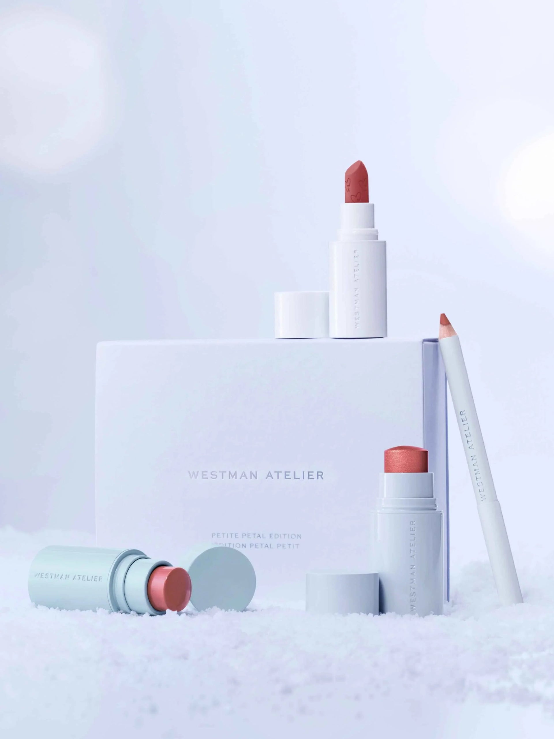 Elegant makeup set featuring rose lipstick, peach blush stick, and soft nude pencil on snowy white backdrop.