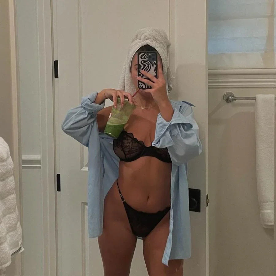 Woman in black lace lingerie and pale blue shirt, hair wrapped in towel, holding green smoothie in luxe bathroom