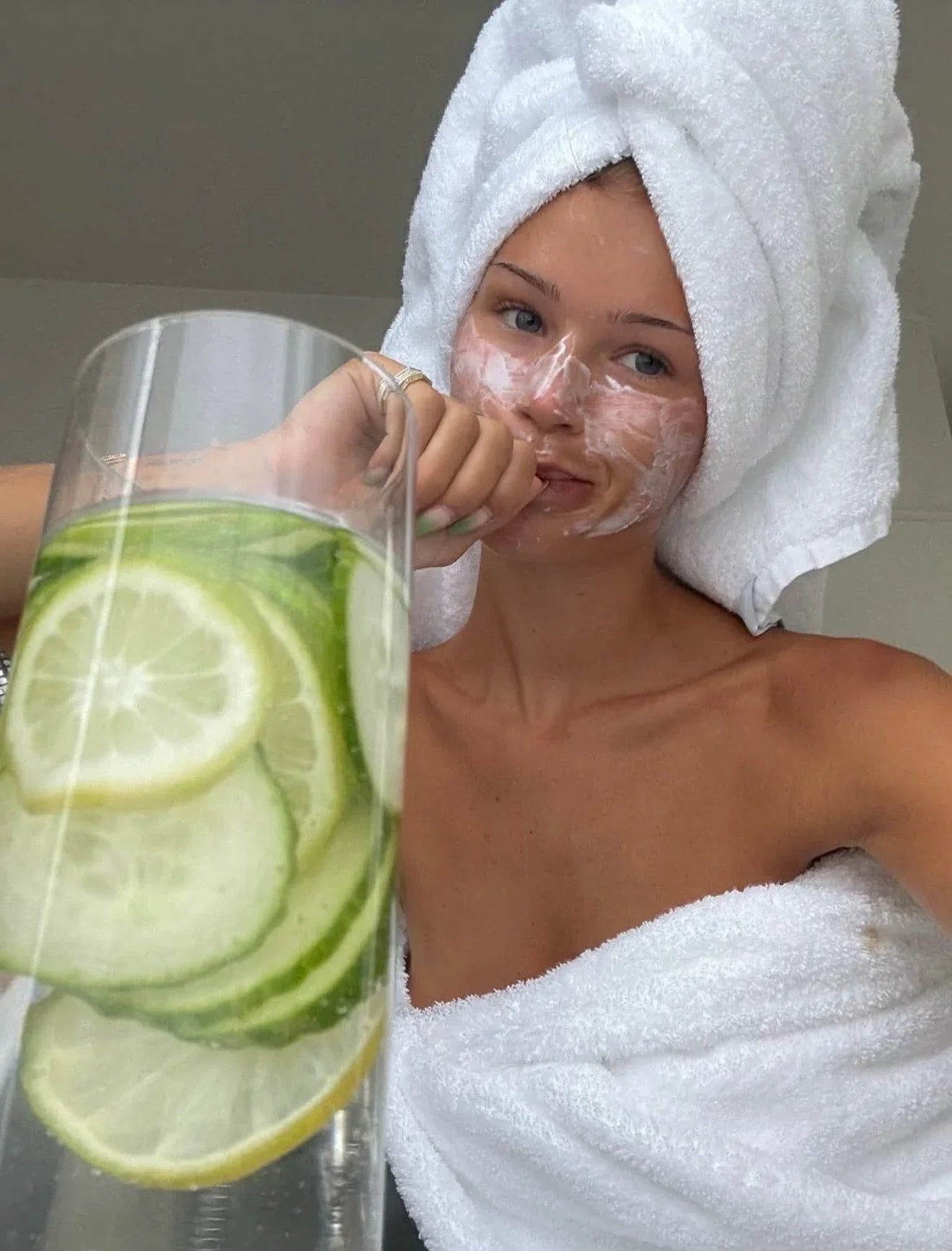 Woman in white towel with a creamy face mask, holding a glass of cucumber lemon water