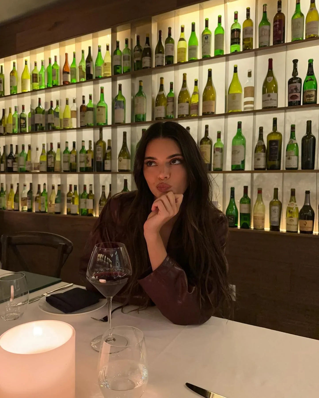 Woman with sleek dark hair in a chic restaurant, wine glass and candle on table, backlit wine bottle wall display