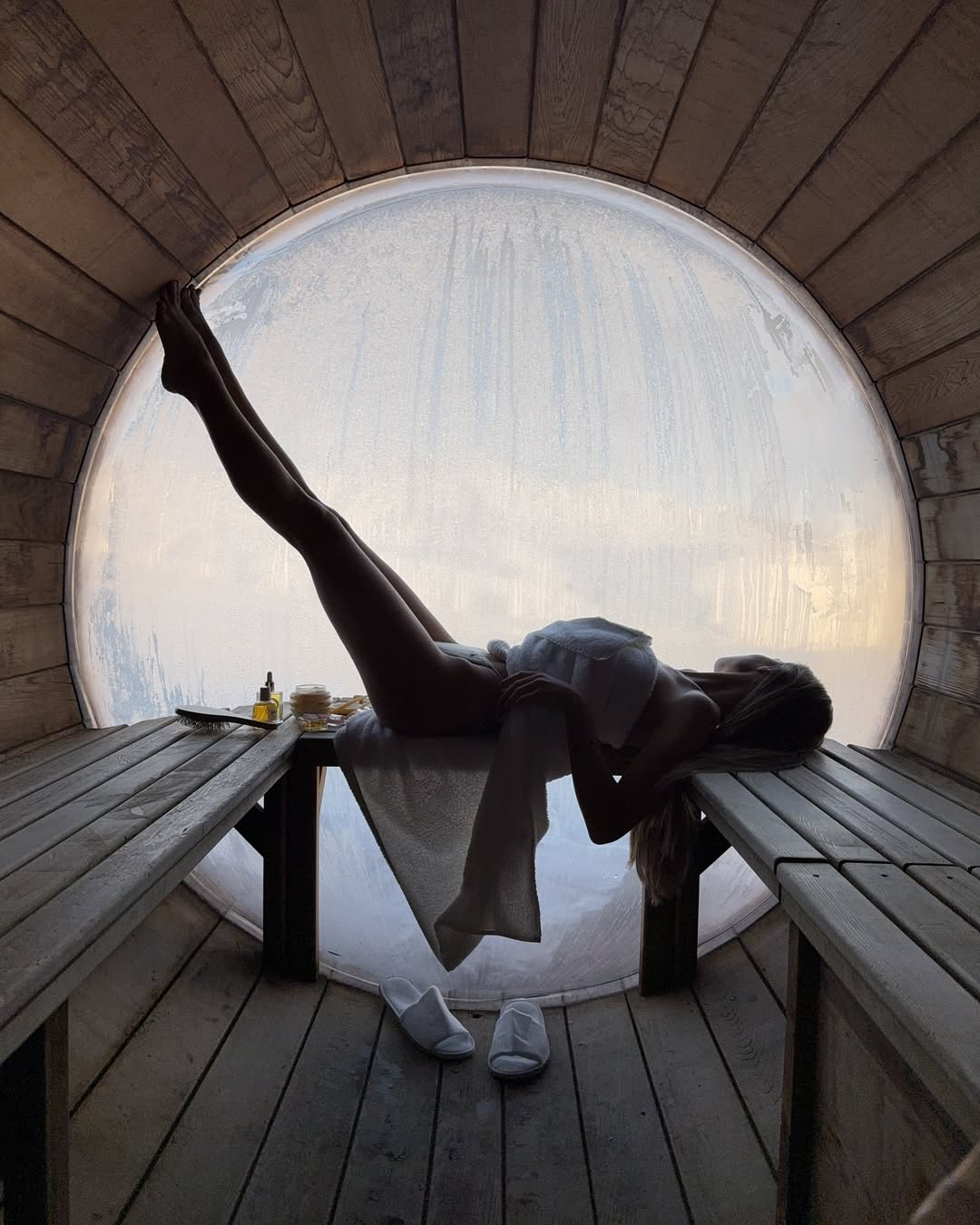Woman relaxing in a wooden sauna with round frosted window, wearing towel and white slippers