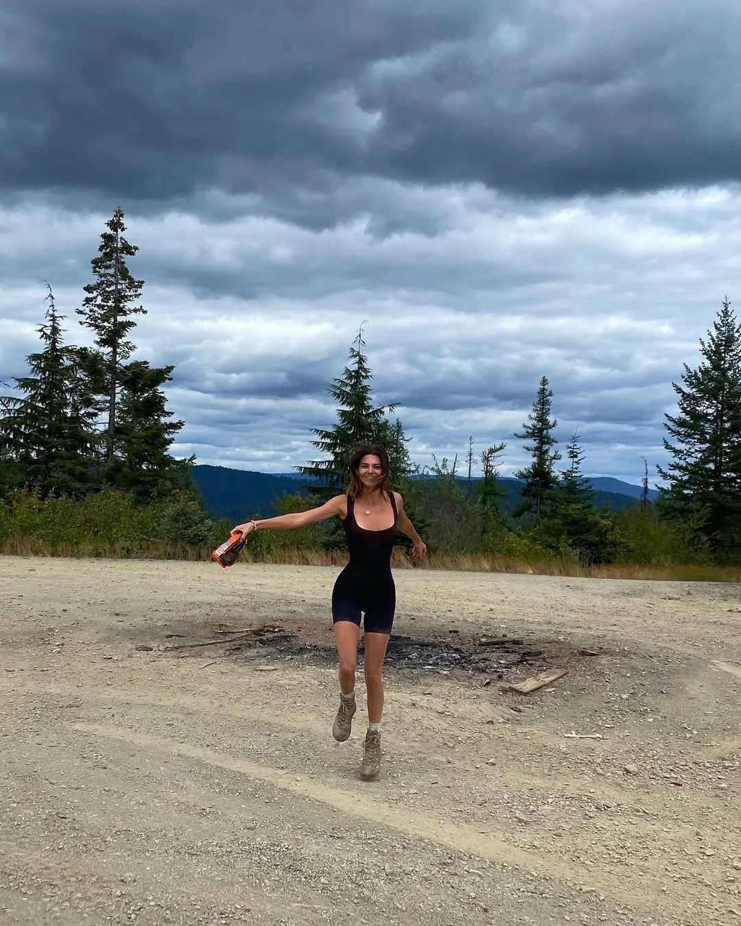 Woman in sleek black athletic romper with dewy skin, outdoors on gravel with pine trees and dramatic cloudy sky.
