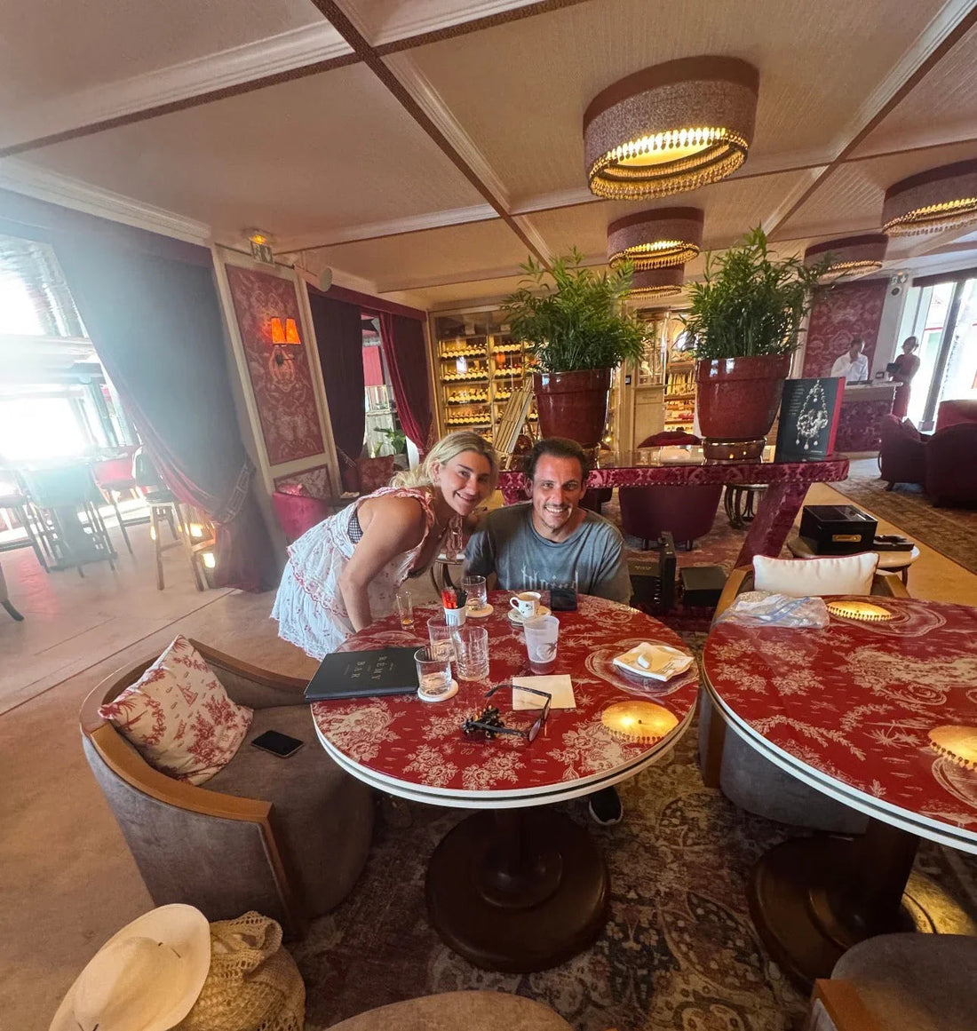 Elegant lounge with red patterned tables, lush green plants, and a couple enjoying drinks
