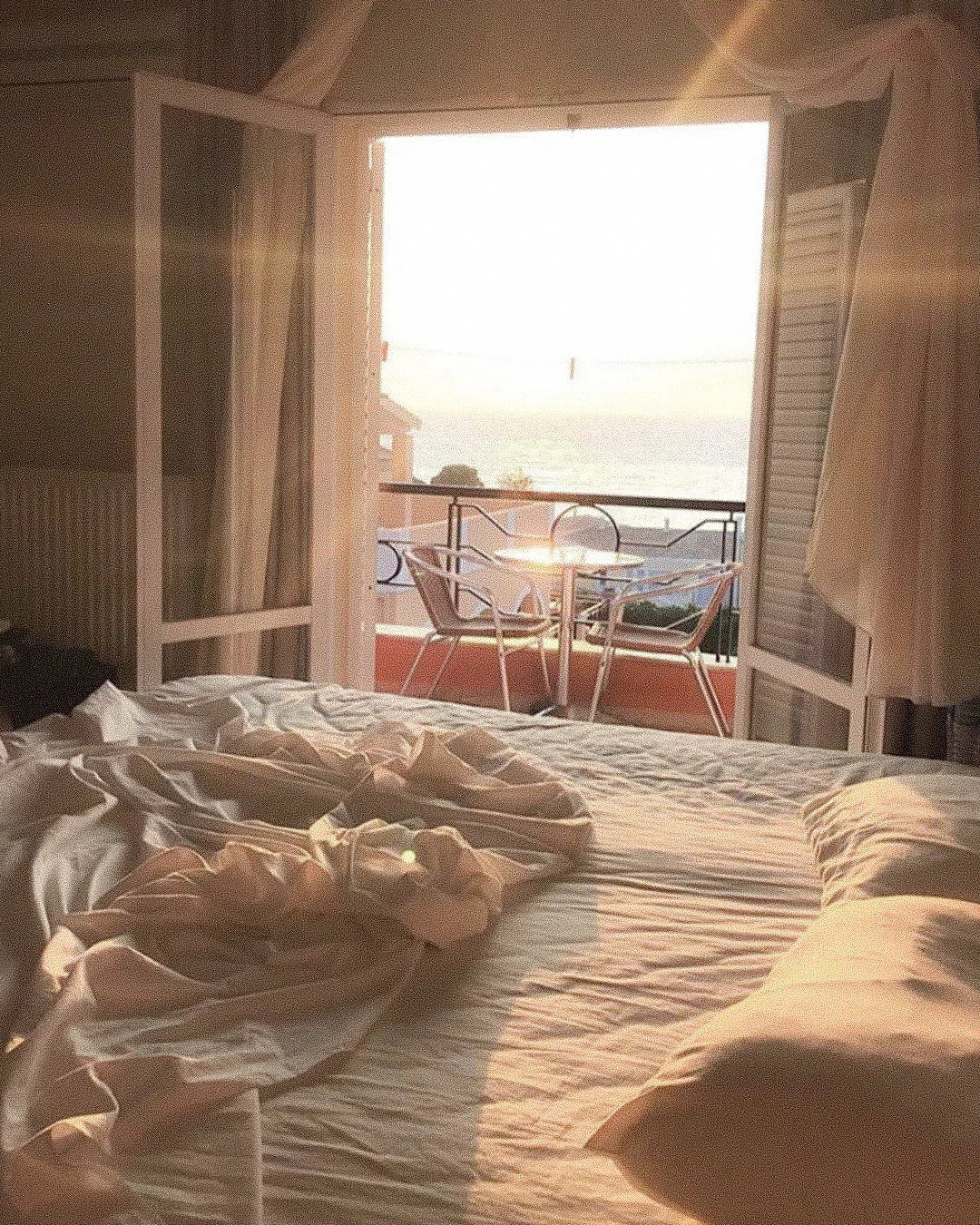 Sunlit bedroom with soft cream bedding, open balcony doors, and ocean view at sunrise.