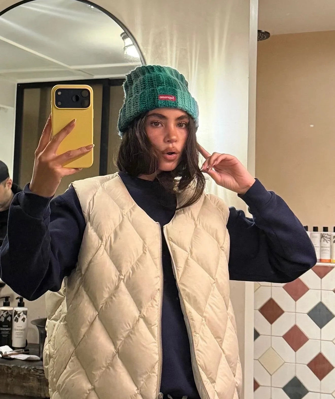 Woman in a quilted beige vest and green knit beanie taking a mirror selfie in a modern bathroom.