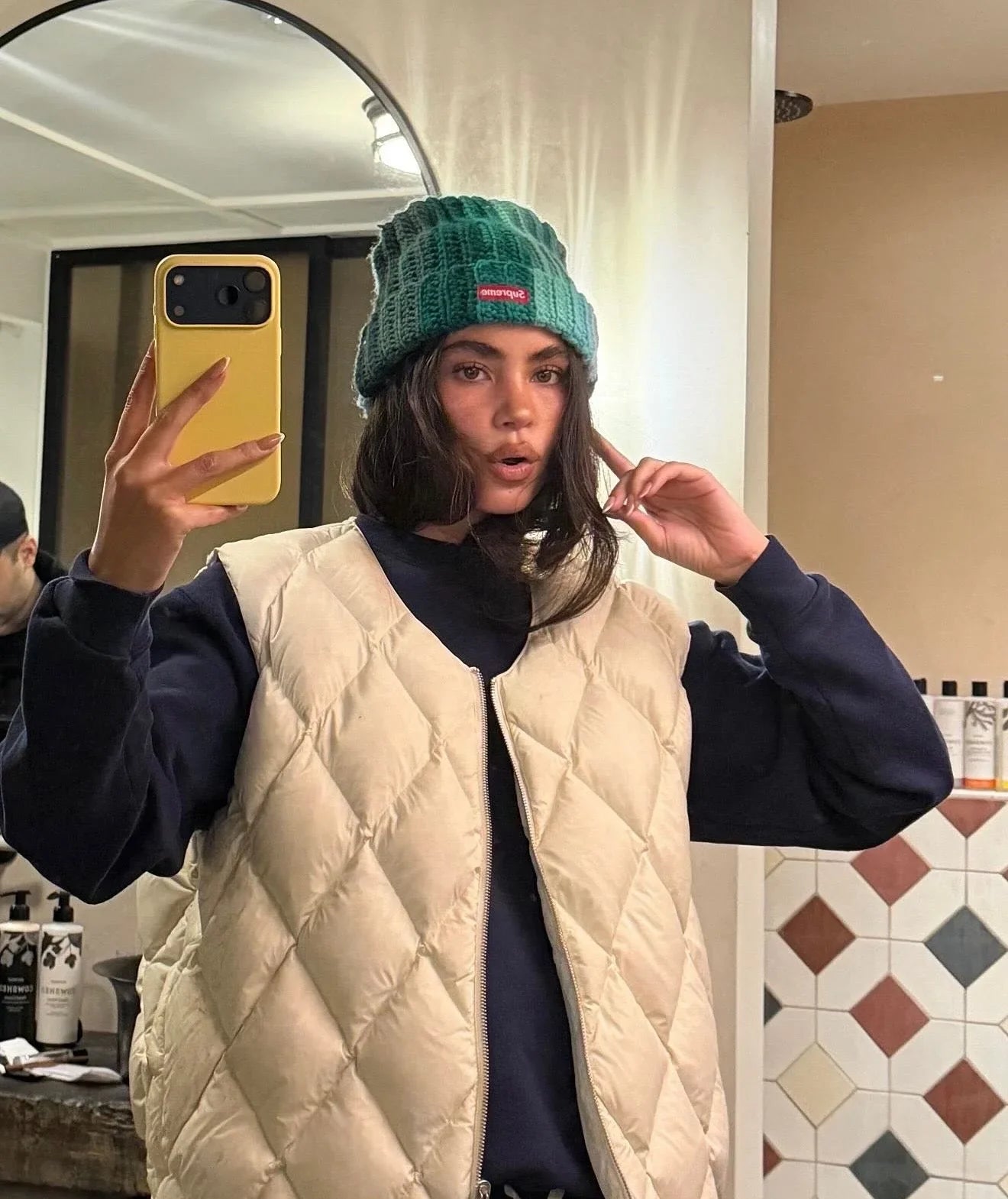 Woman in a quilted beige vest and green knit beanie taking a mirror selfie in a modern bathroom.