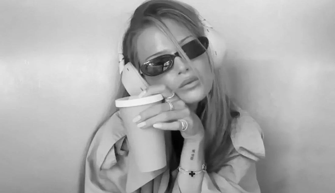 Chic woman in sunglasses and headphones, with glossy lips and dewy makeup, holding coffee cup.