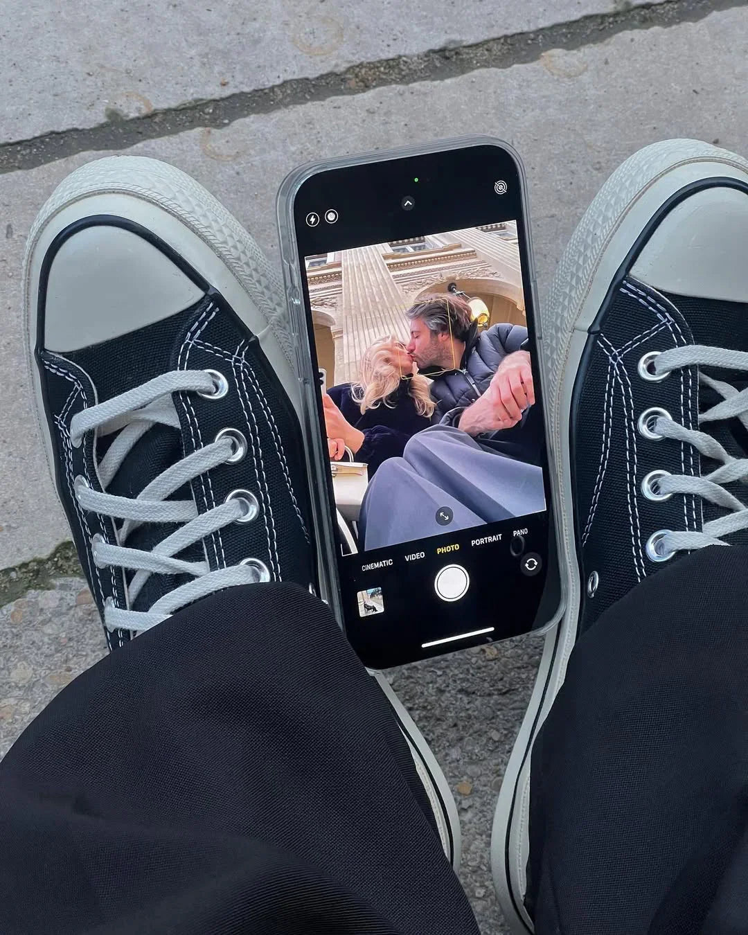 Black canvas sneakers framing a smartphone capturing a couple embracing outdoors.