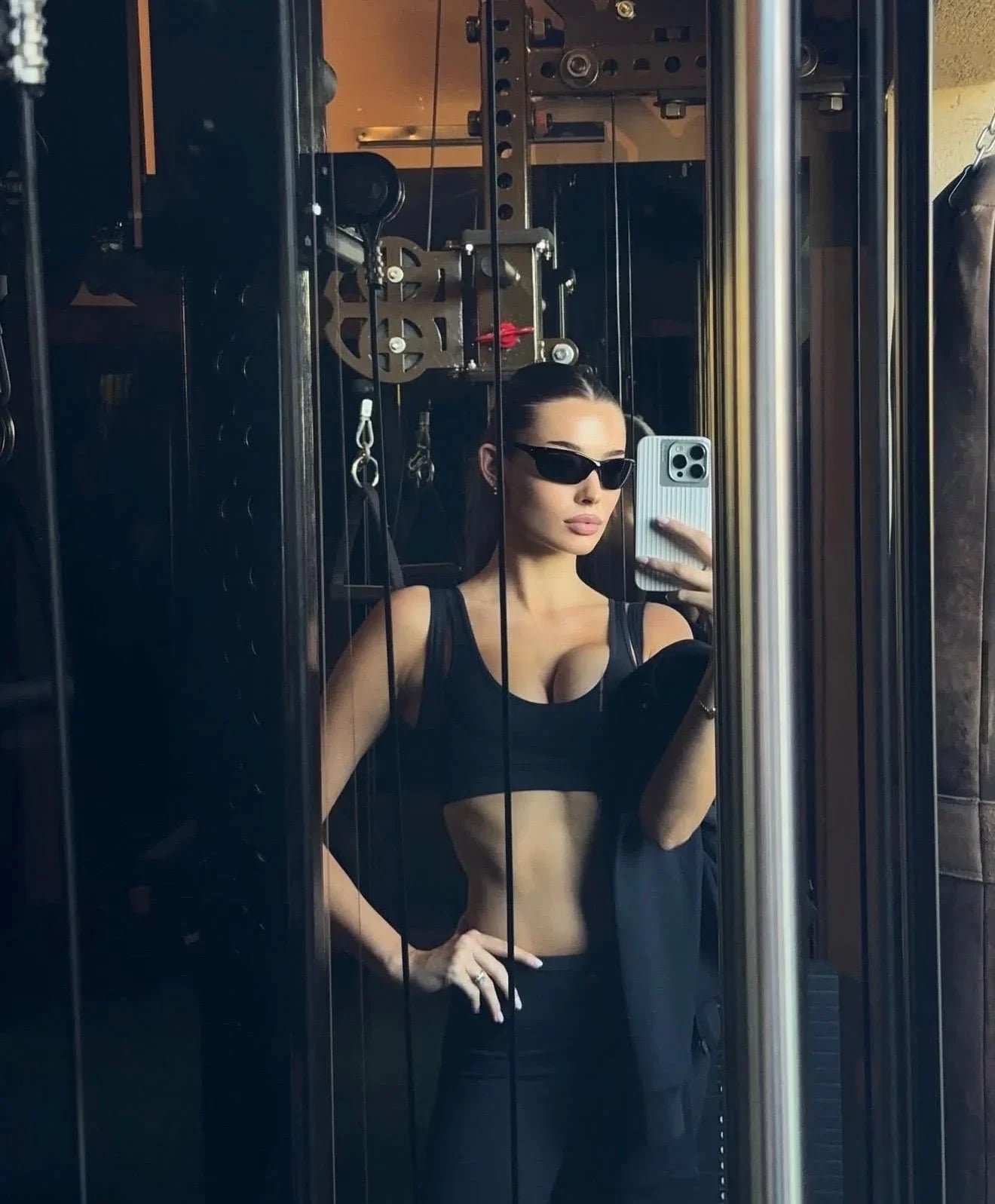 Woman in sleek black activewear and sunglasses taking a mirror selfie in a modern gym setting.