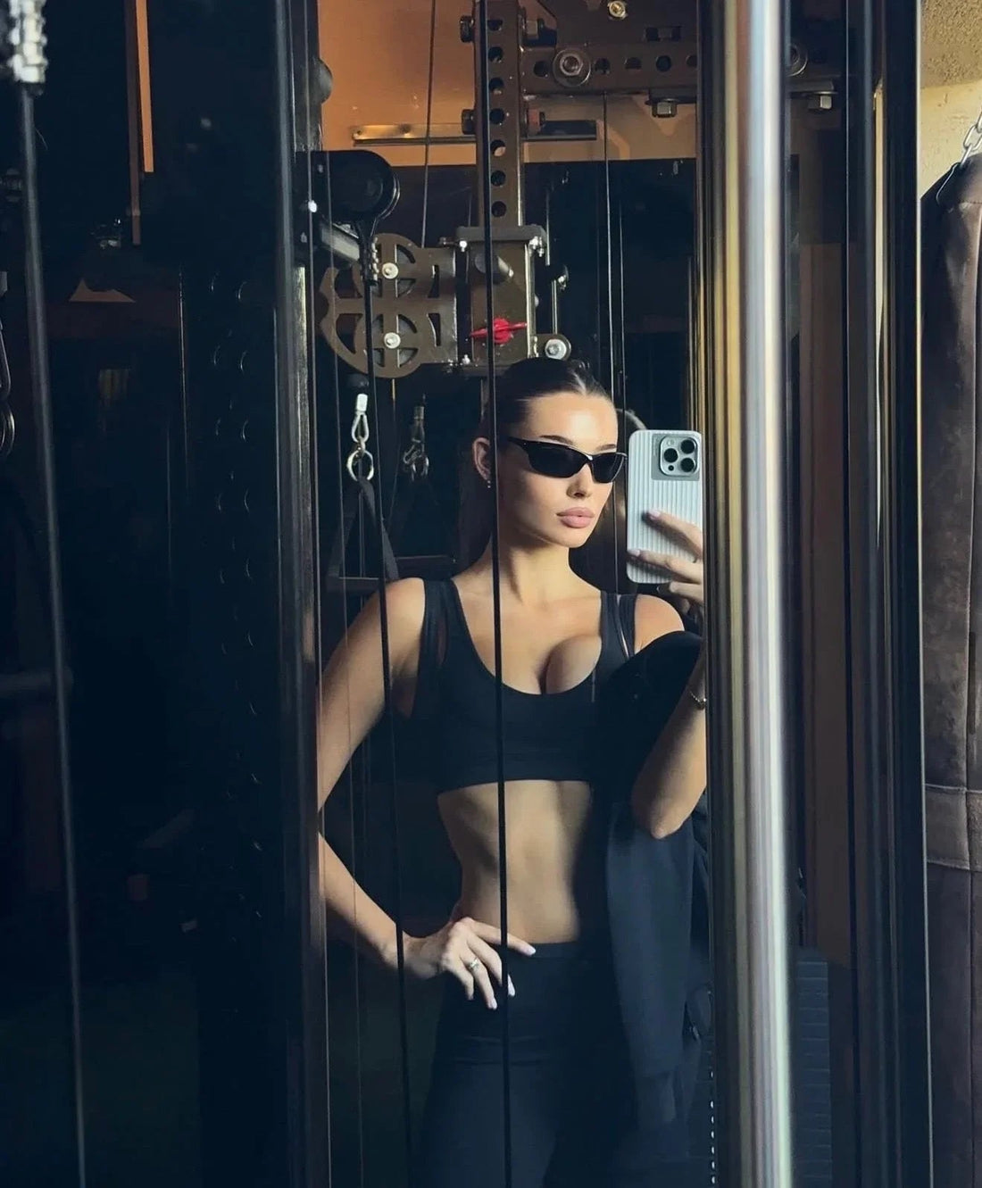 Woman in sleek black workout set and sunglasses, taking a mirror selfie in a modern gym.