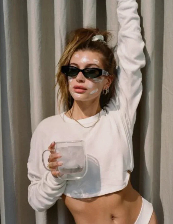 Woman with glowing skin and visible cream spots, wearing sunglasses and white loungewear, holding a clear glass of ice.