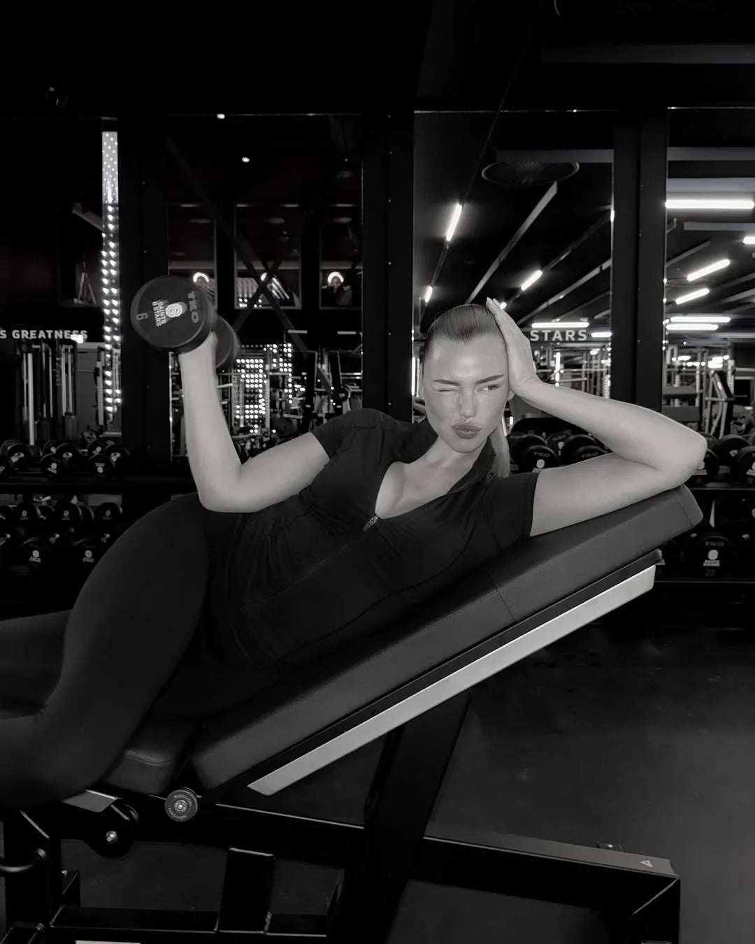 Woman in sleek black activewear lifting dumbbell on gym bench, modern gym background, Wylde Grey