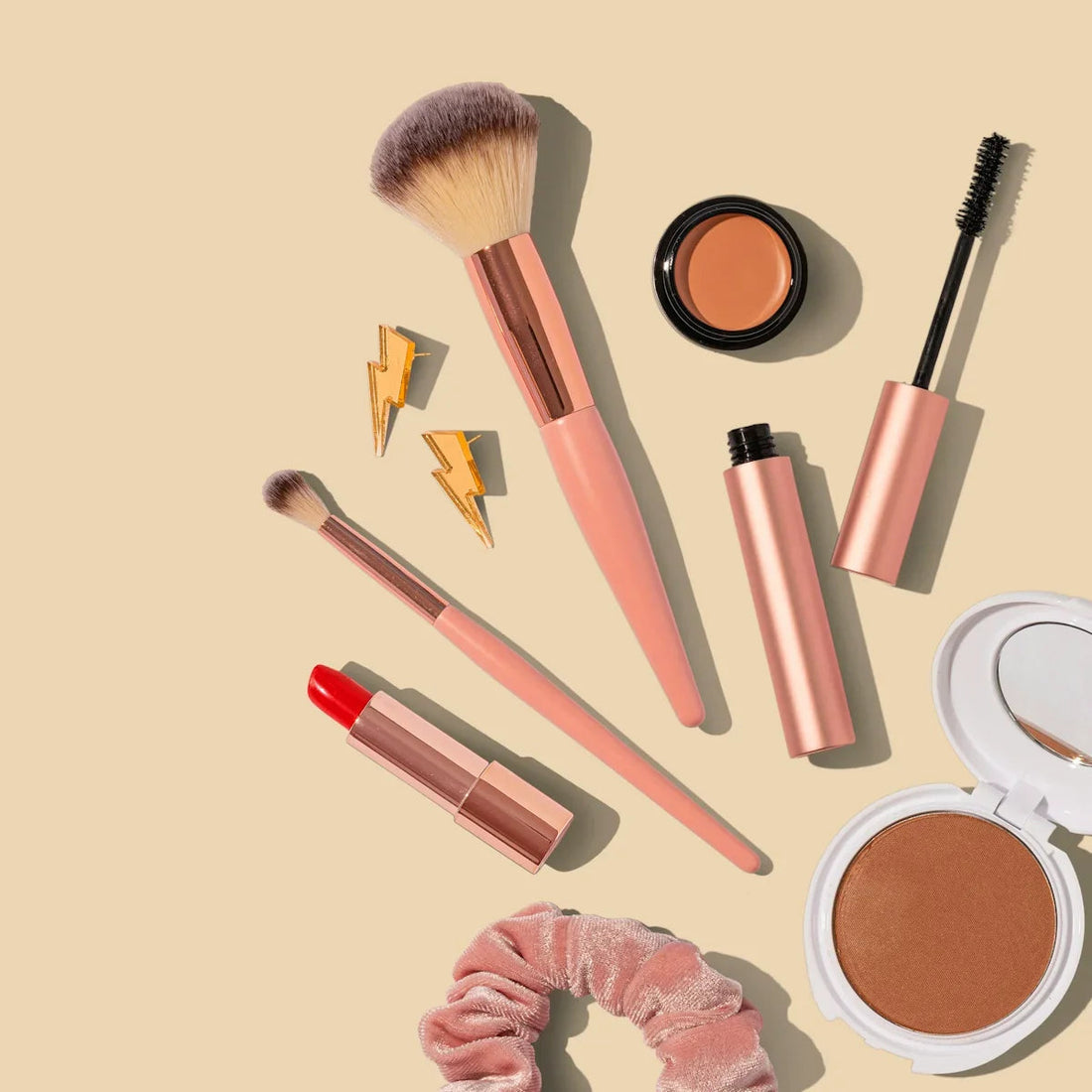 Luxury makeup flatlay with rose gold brushes, bold red lipstick, creamy bronzer, matte compact, velvet scrunchie, and gold lightning bolt earrings on a beige background.