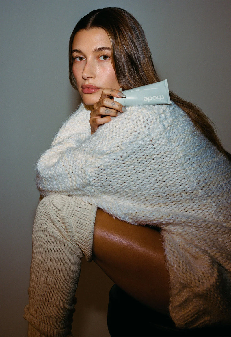 Woman in chunky white sweater and cream leg warmers holds Node skincare tube, showcasing dewy skin.