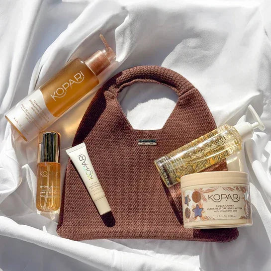 Luxurious skincare and body care products with golden oils and creamy textures arranged around a textured brown handbag on white satin.