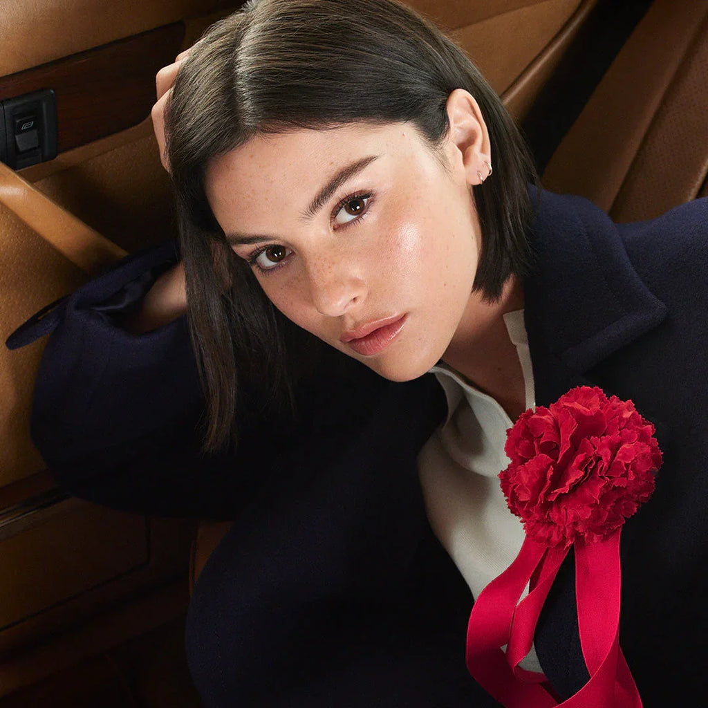 Woman with glowing skin, sleek dark hair, navy coat, red flower pin, luxury beauty look