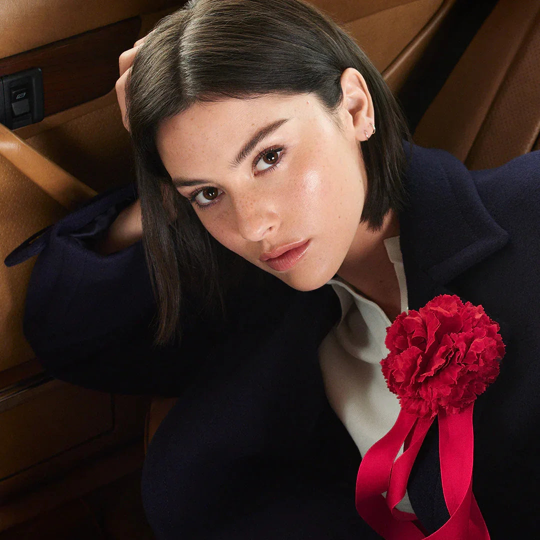 Woman with glowing skin, sleek dark hair, navy coat, red flower pin, luxury beauty look