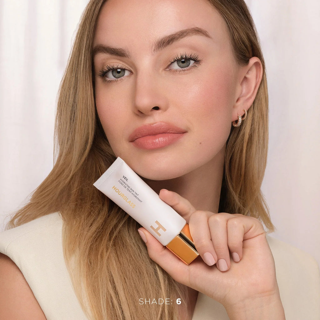 Model with glowing skin holding Hourglass Veil foundation, shade 6, luxurious soft finish