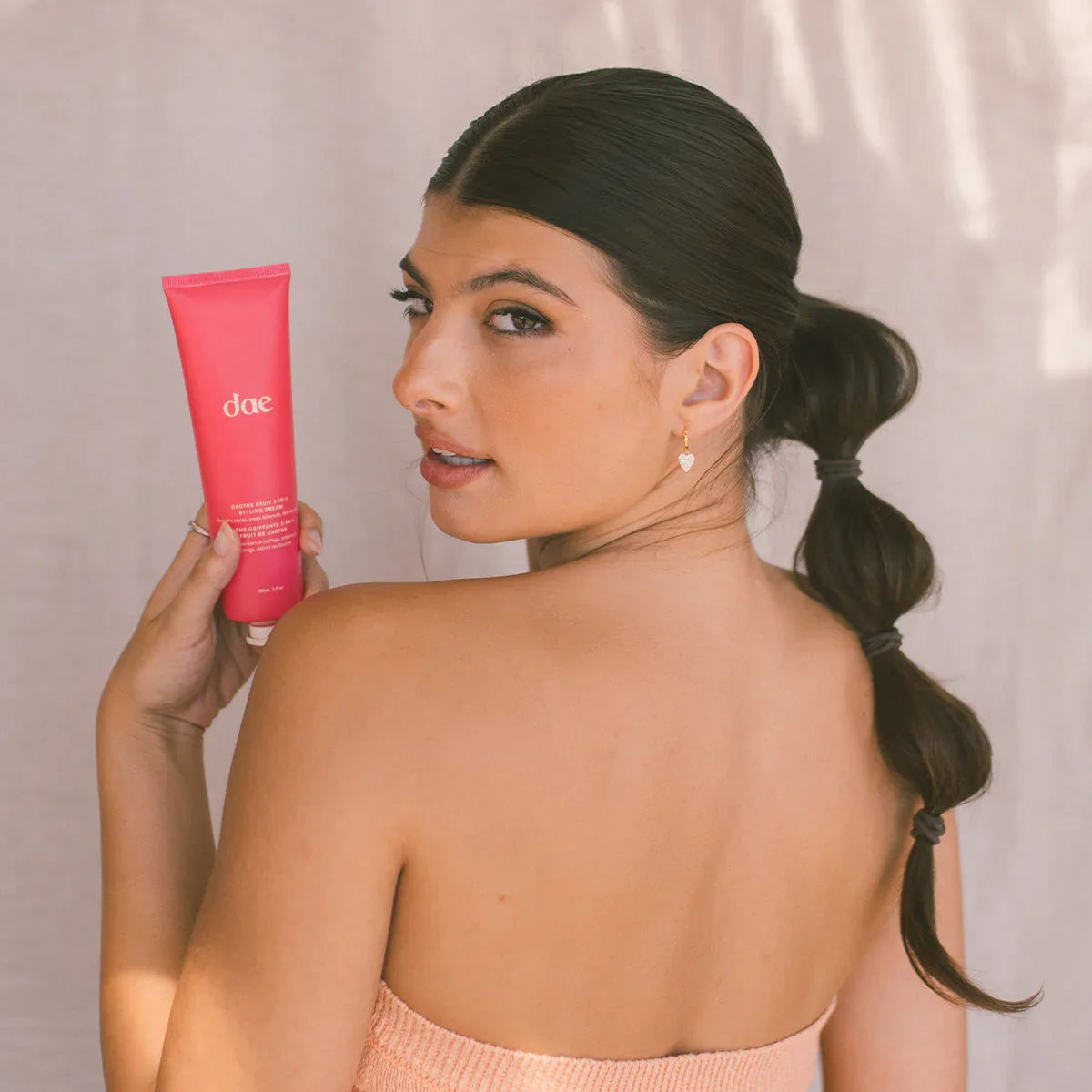Woman with sleek bubble ponytail holding Dae pink beauty tube, glowing skin, soft neutral backdrop