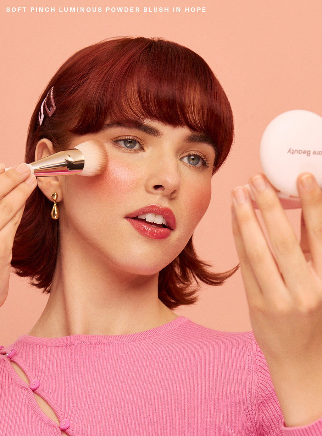 Hope| A model with short red hair wearing a pink sweater applies Rare Beauty's Soft Pinch Luminous Powder Blush using the Rare Beauty Soft Pinch Blush Brush.