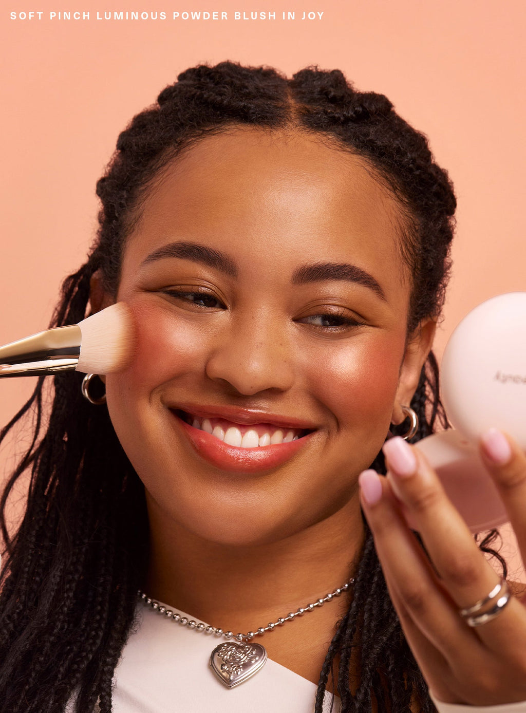 Joy| A model smiles and applies Rare Beauty's Soft Pinch Luminous Powder Blush in the shade Joy.