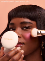 Truth| A model with short bangs looks into the camera and applies Rare Beauty's Soft Pinch Luminous Powder Blush using the Rare Beauty Soft Pinch Blush Brush.