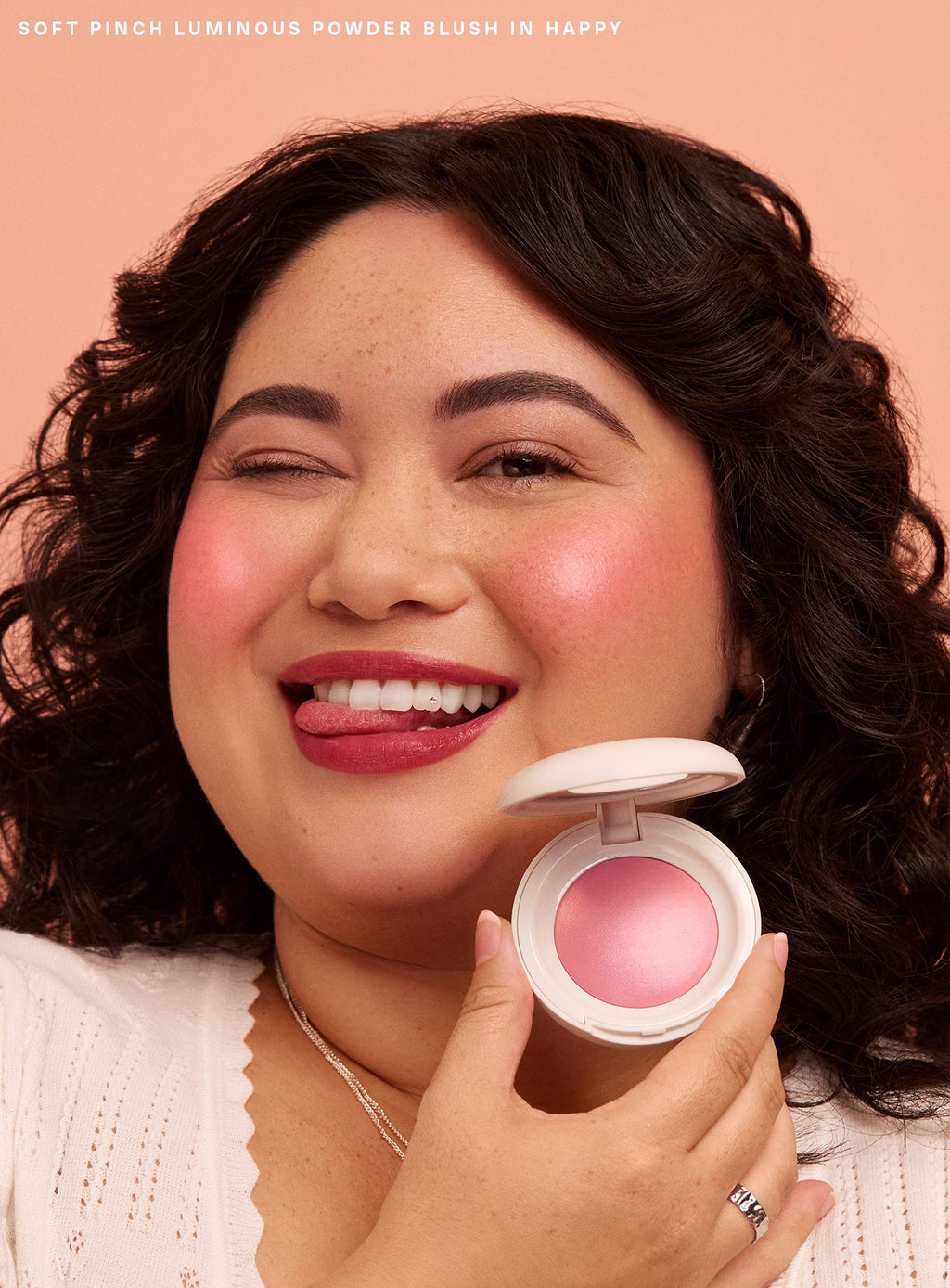 Happy| A model with curly hair sticks her tongue out and winks while holding Rare Beauty's Soft Pinch Luminous Powder Blush in the shade Happy.