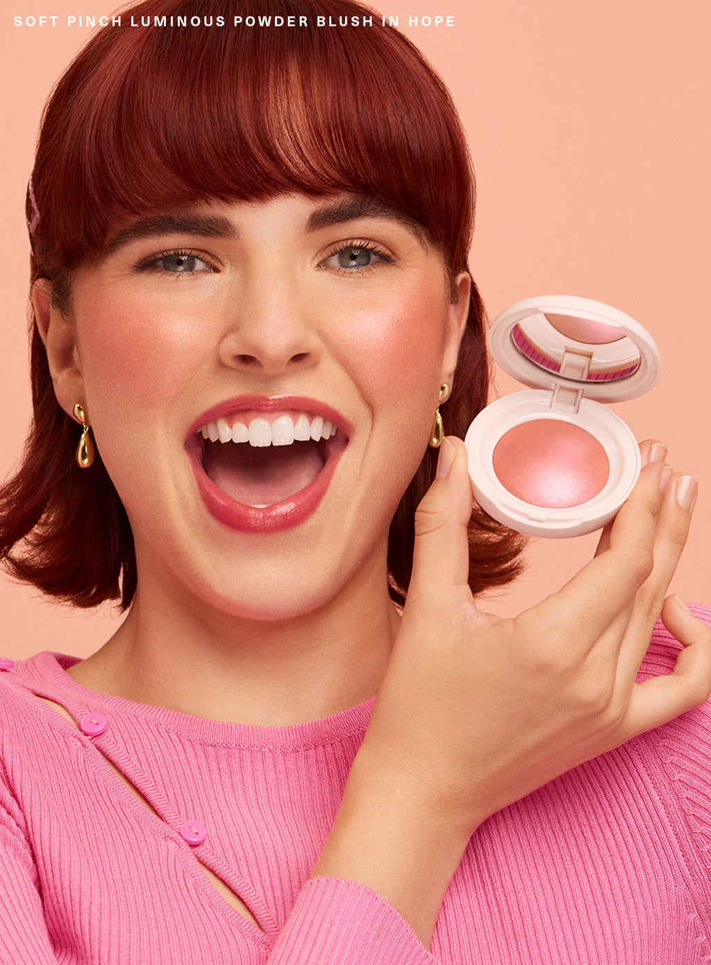 Hope| A model with short red hair smiles and excitedly holds up Rare Beauty's Soft Pinch Luminous Powder Blush.