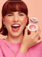 Hope| A model with short red hair smiles and excitedly holds up Rare Beauty's Soft Pinch Luminous Powder Blush.