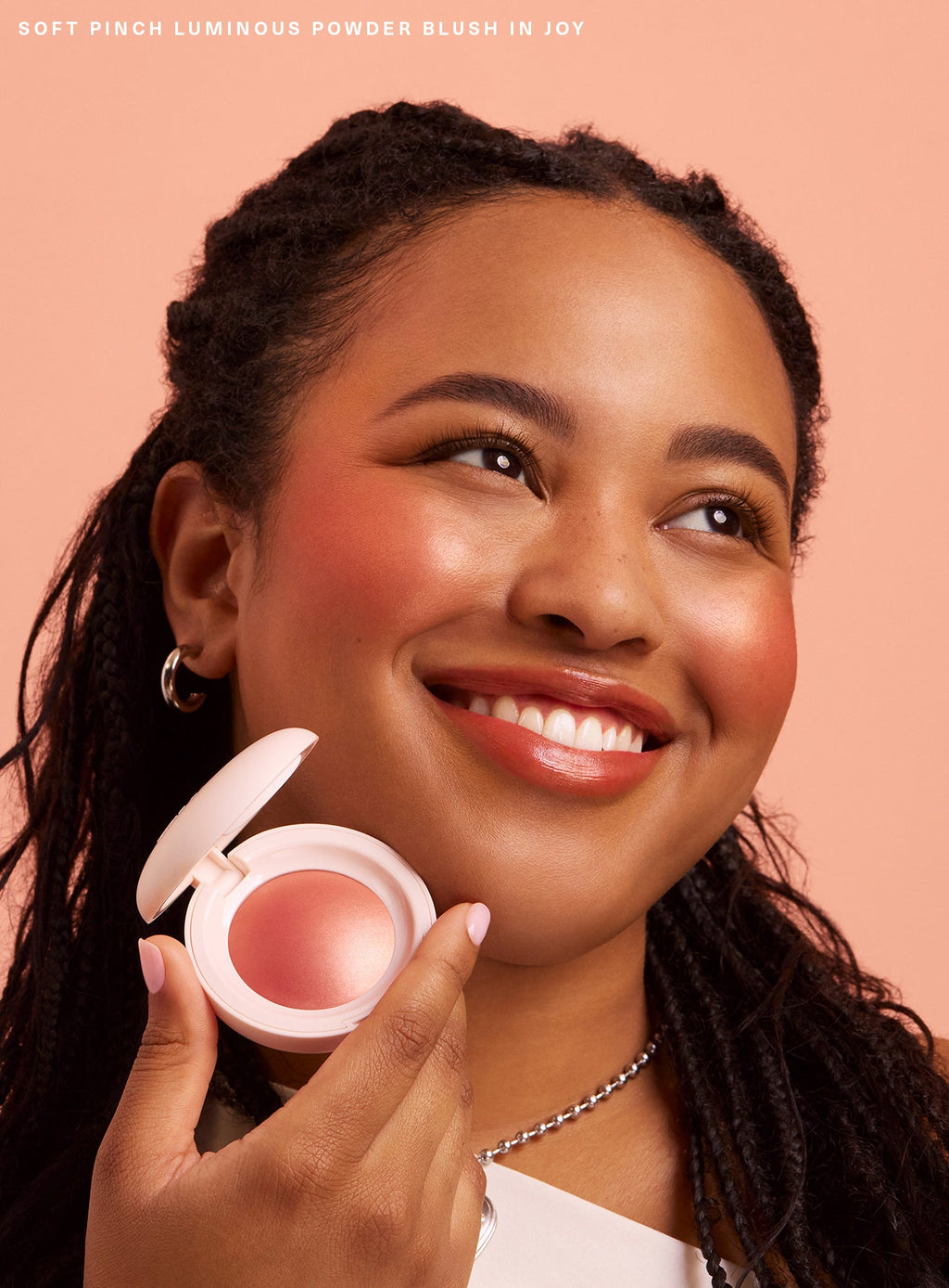 Joy| A model smiles and holds up Rare Beauty's Soft Pinch Luminous Powder Blush in the shade Joy while looking up and off to the right.