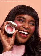 Truth| A model smiles and holds up Rare Beauty's Soft Pinch Luminous Powder Blush in the shade Truth while looking up and off to the right.