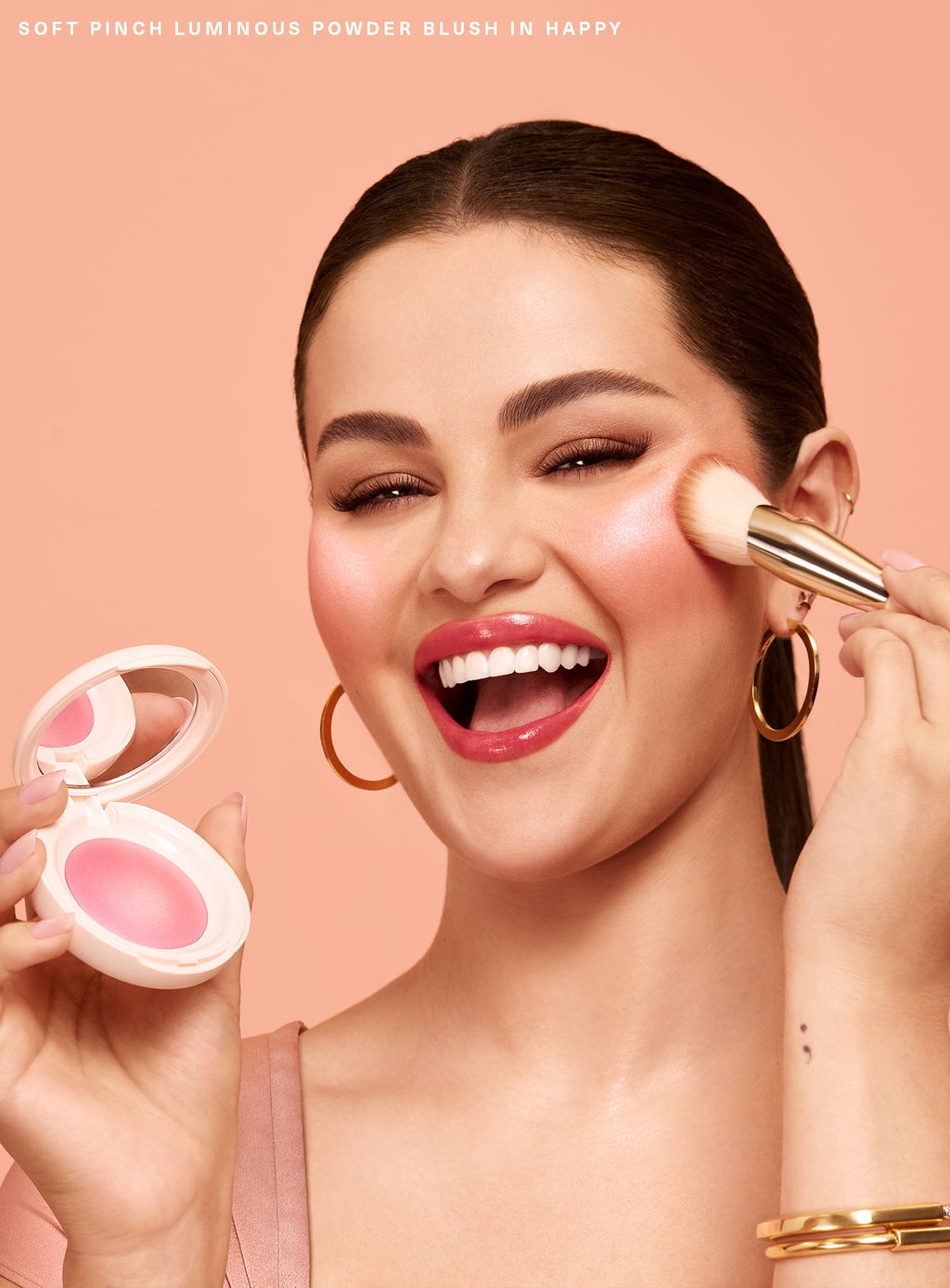 Happy| Selena Gomez smiles excitedly while applying Rare Beauty's Soft Pinch Luminous Powder Blush in the shade Happy with the Rare Beauty Soft Pinch Blush Brush.