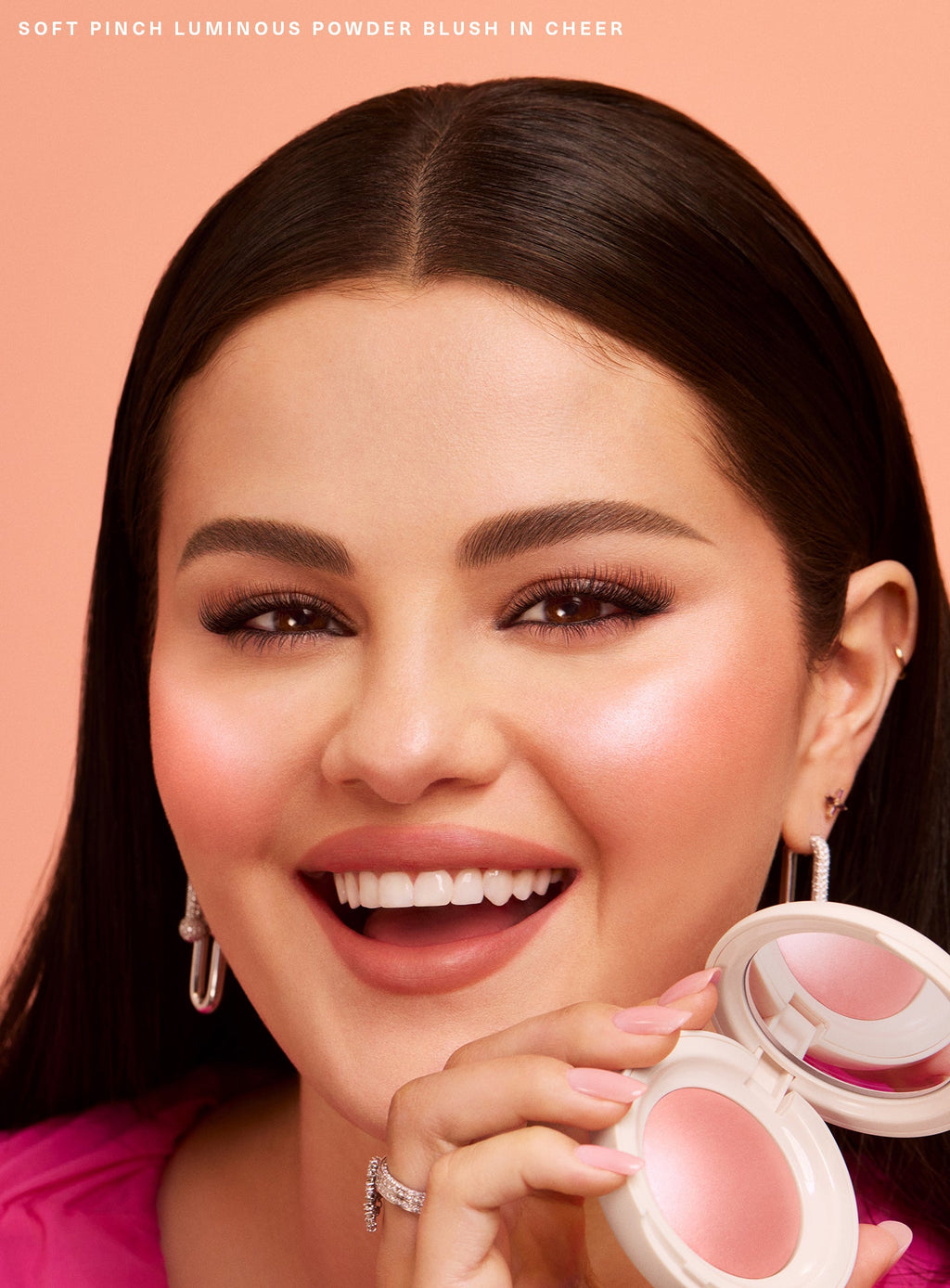 Cheer| Selena Gomez holds Rare Beauty's Soft Pinch Luminous Powder Blush in the shade Cheer with smoothed down straight hair and a happy expression. 