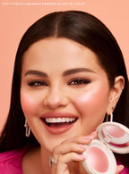 Cheer| Selena Gomez holds Rare Beauty's Soft Pinch Luminous Powder Blush in the shade Cheer with smoothed down straight hair and a happy expression. 