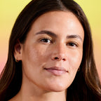 Close-up of woman with natural glowy skin, subtle freckles, and gold hoop earrings, yellow background