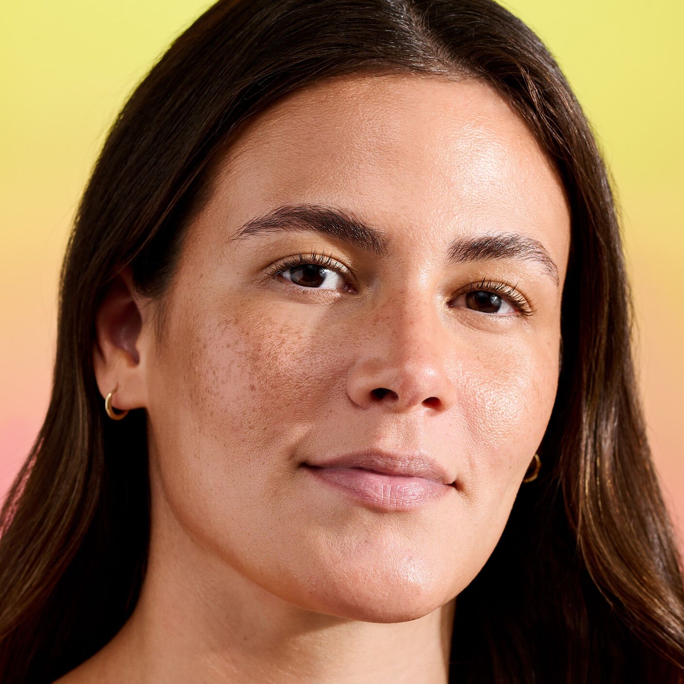 Close-up of woman with natural glowy skin, subtle freckles, and gold hoop earrings, yellow background