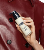 Hand with glossy nude nails holding Chris McMillan Styling Balm against burgundy leather jacket