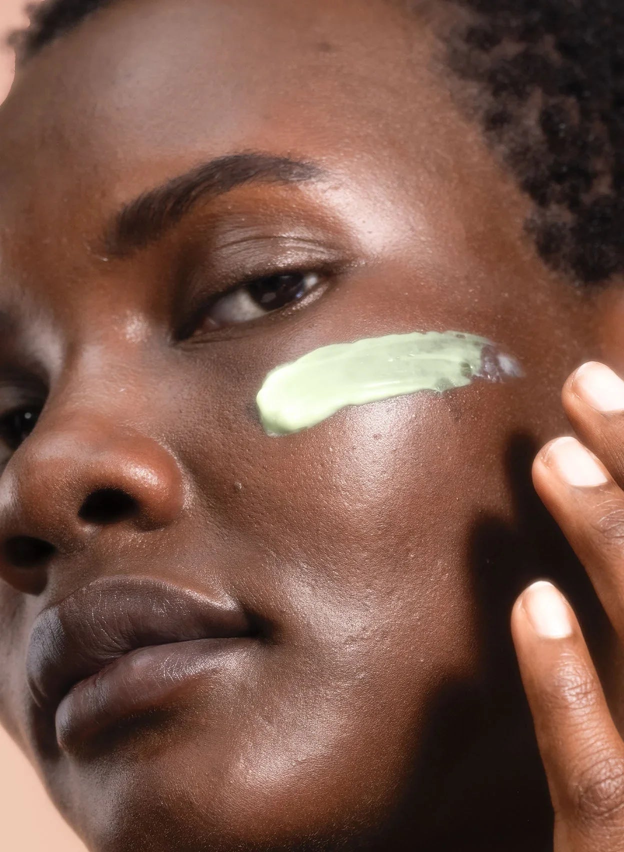 Model with glowing skin applying creamy green skincare product on cheek