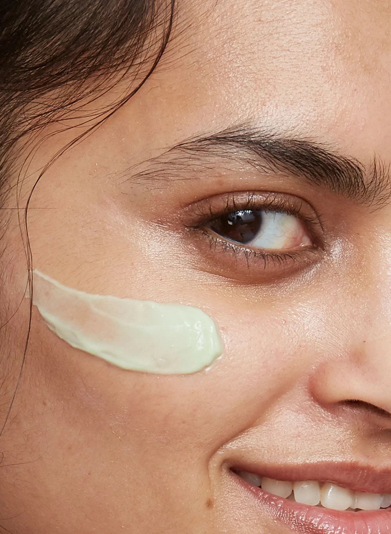 Close-up of woman with dewy skin applying creamy, light green skincare product on cheek