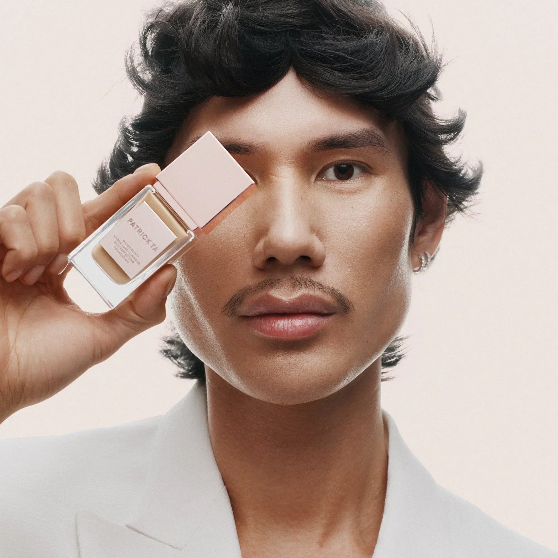 Model in a white blazer holds Patrick Ta Luxe Foundation bottle, creamy beige shade, radiant finish