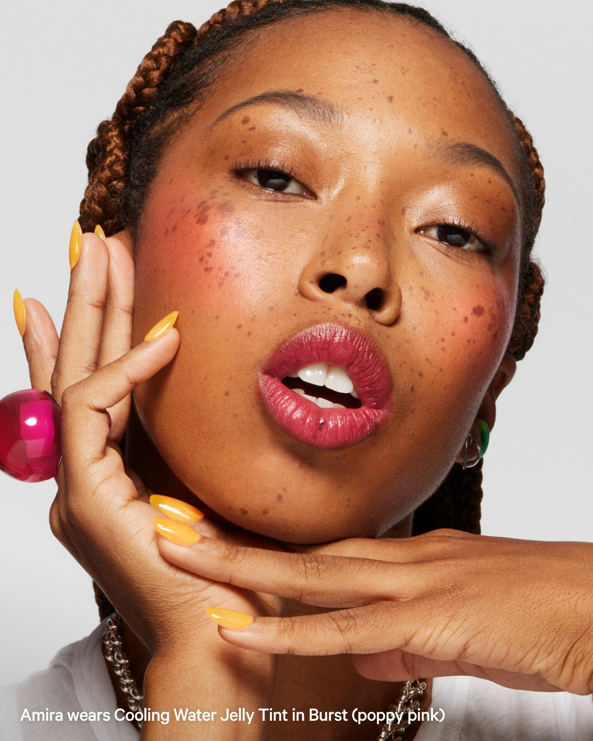 Woman with braided hair, bold pink lips, glossy cheeks, yellow nails, and a pink ring close-up