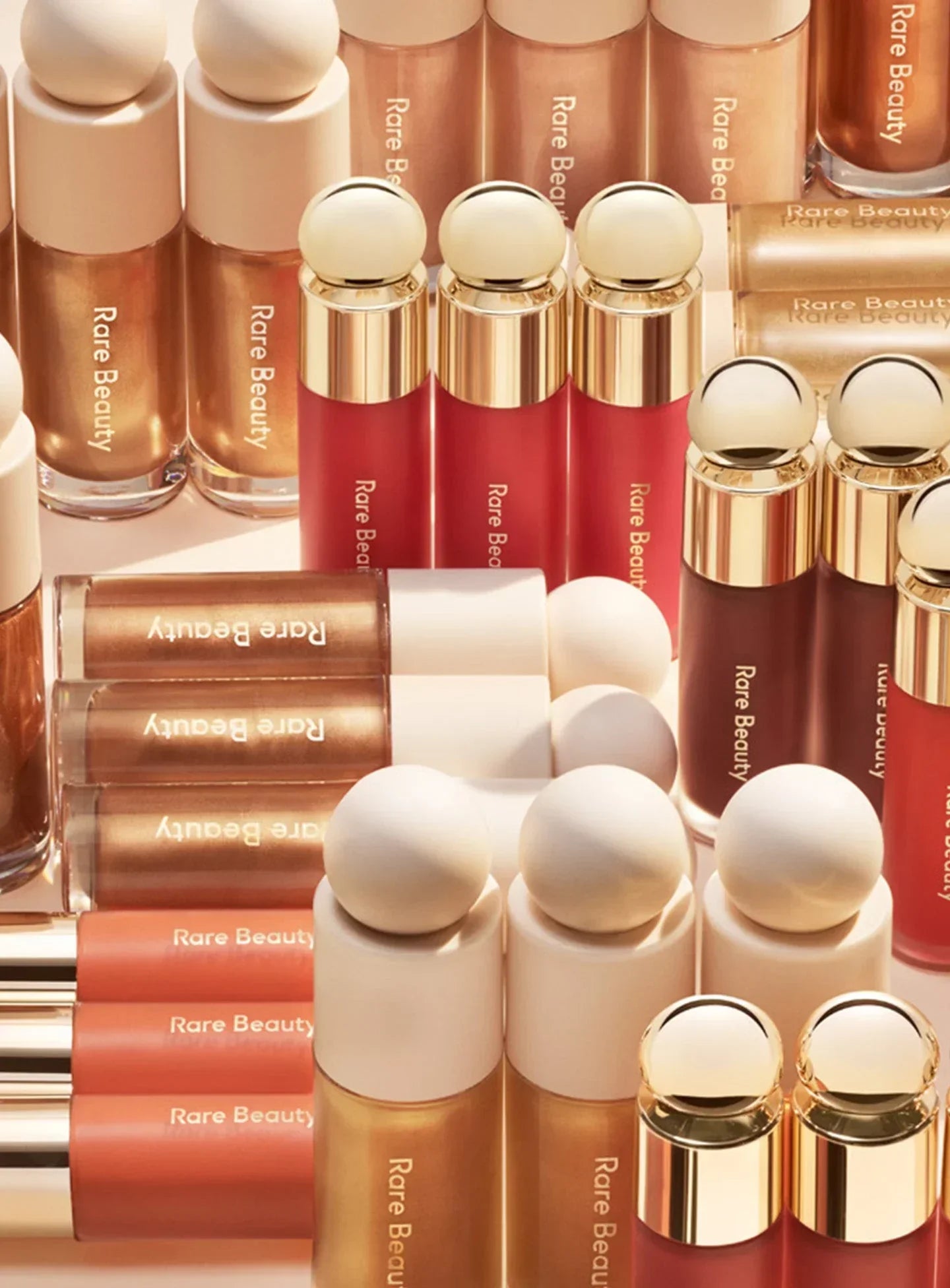 Assorted containers of Rare Beauty's liquid luminizer, blush, and contour, some laying and some standing up.