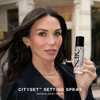 Woman with dewy complexion holding Saie Cityset Setting Spray, elegant outdoor setting