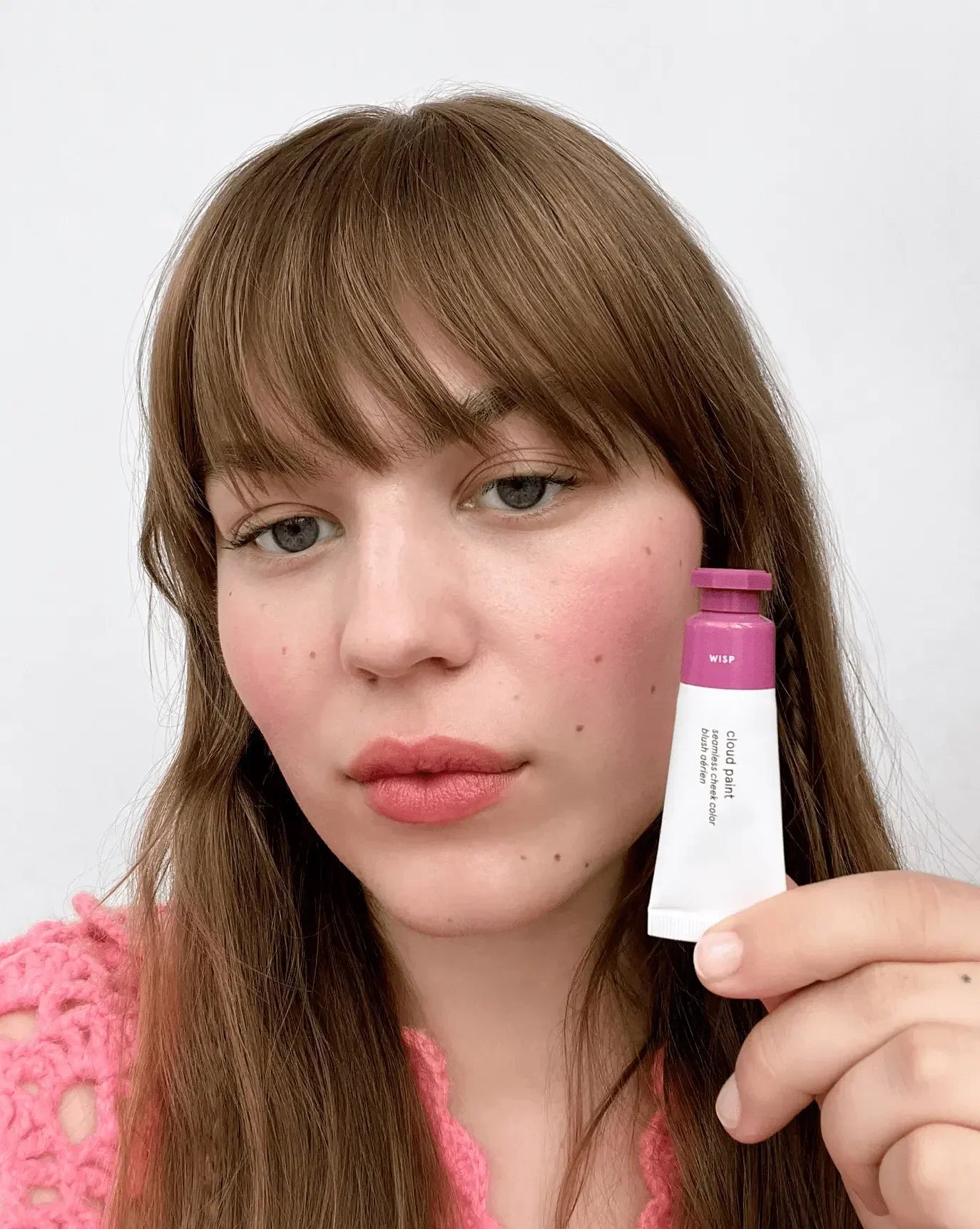 Woman holding pink cream blush tube with soft, dewy blush on her cheeks.