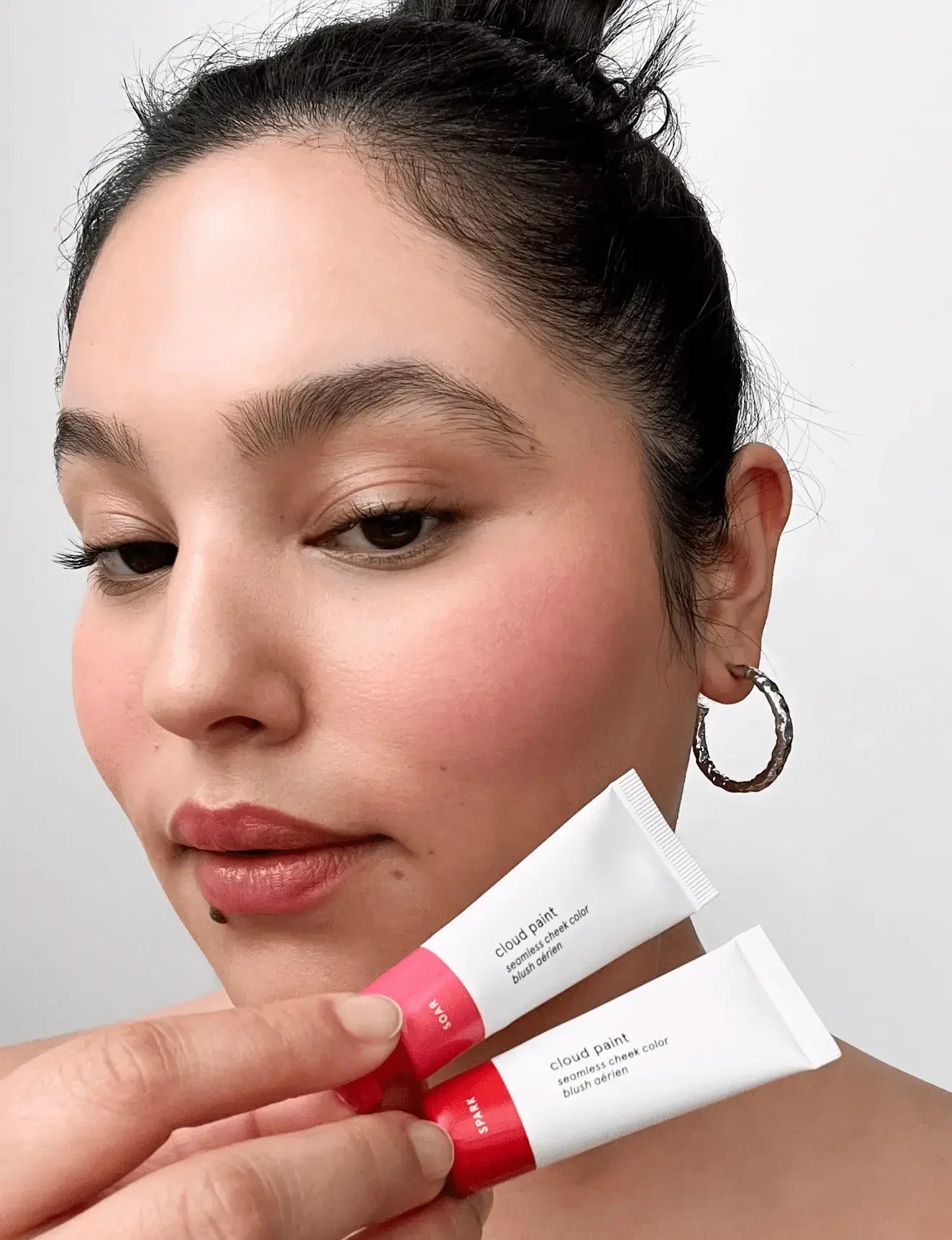 Woman with radiant, rosy blush and dewy finish holding two Cloud Paint cheek color tubes.