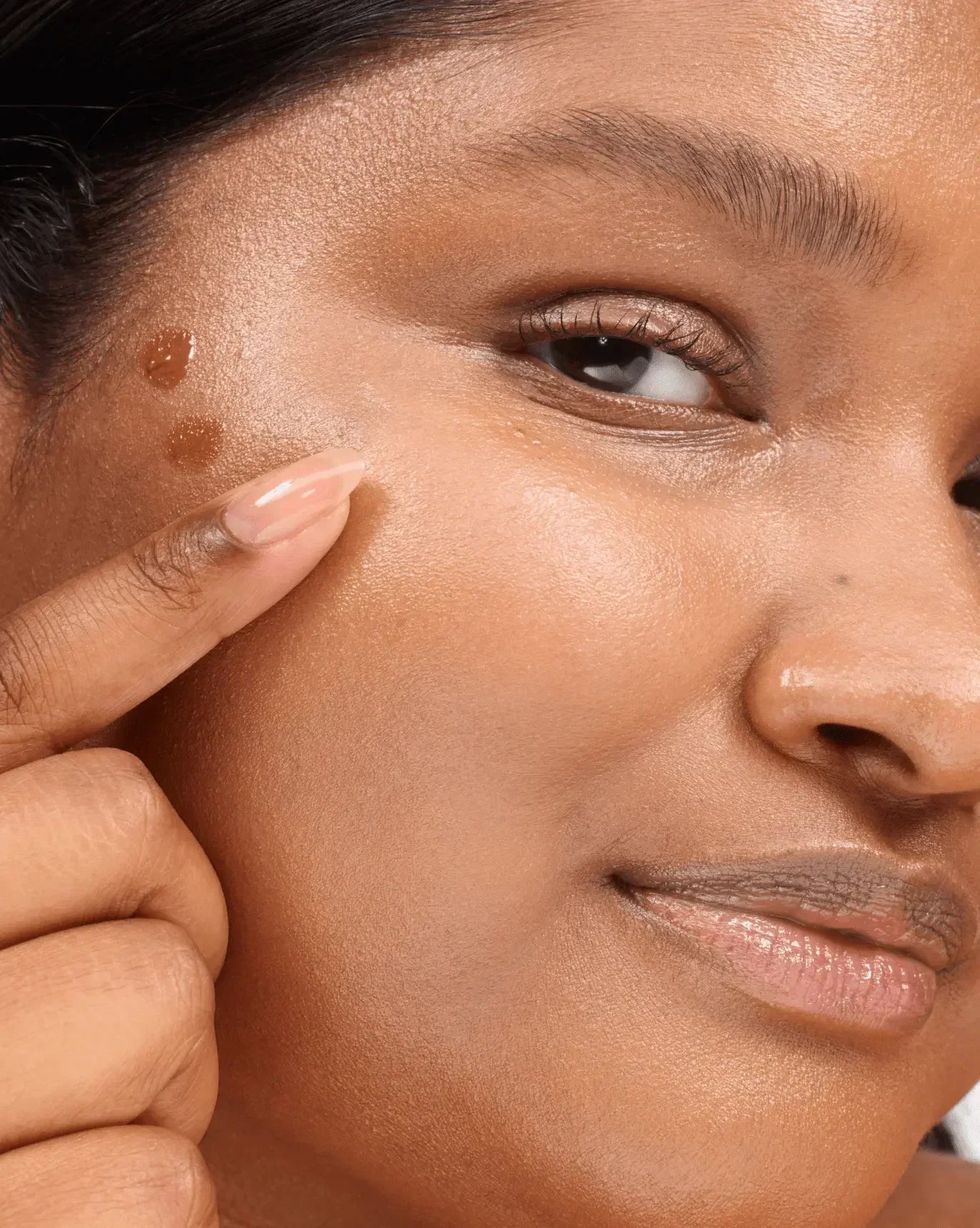 Close-up of a woman applying creamy brown liquid blush with a dewy finish to her cheek.