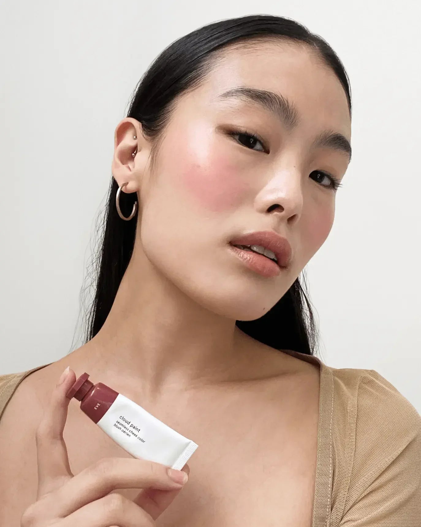 Model with radiant pink blush and dewy finish, holding creamy liquid blush tube