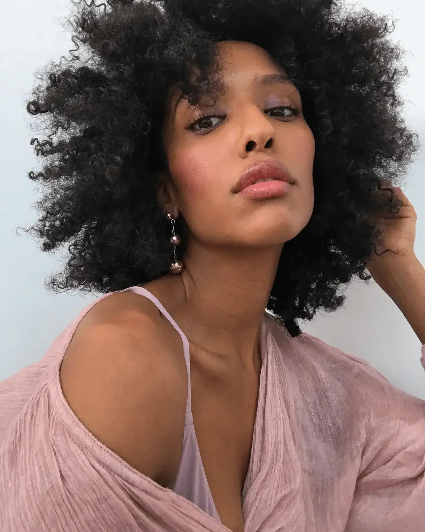 Woman with voluminous natural curls, dewy blush and rosy pink makeup, wearing a mauve top.