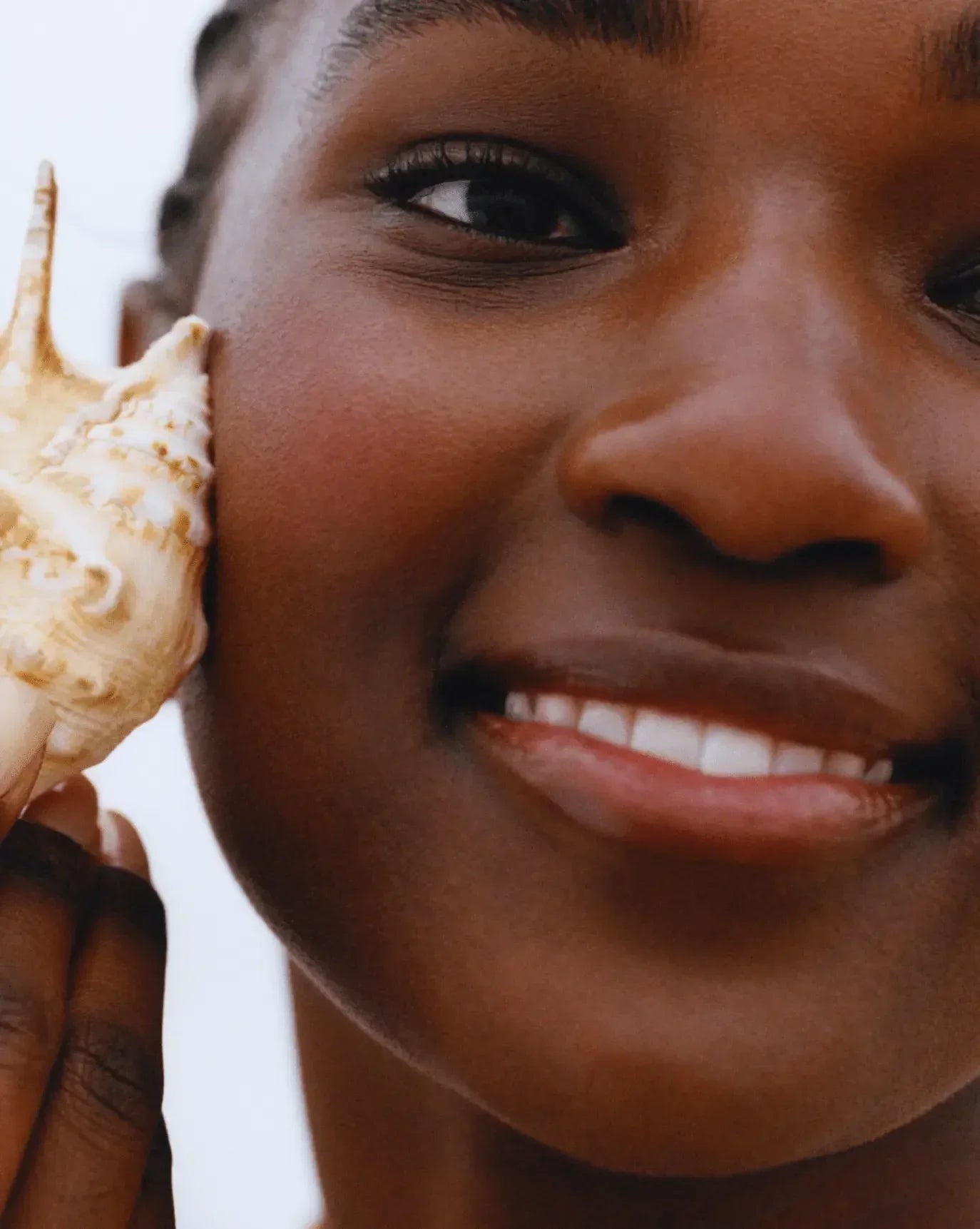 Woman with radiant, dewy skin and soft pink blush, holding a seashell to her cheek